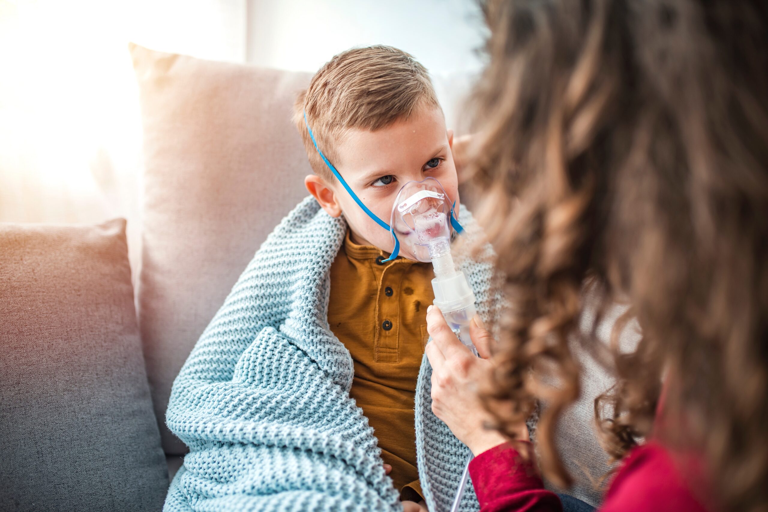 Mukowiscydoza - siedzący na kanapie owinięty kocem chłopiec z inhalatorem na buzi